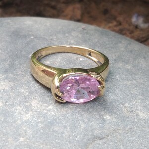 May include: A gold ring featuring a large, oval-shaped, light pink gemstone. The ring's band is thick and has a design that resembles two hands holding the gemstone.