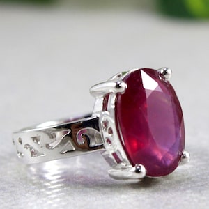 以下が含まれることがあります： 楕円形のルビーの宝石が付いた銀の指輪。指輪は繊細で複雑なデザインです。