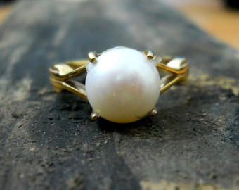 May include: A gold ring with a large, round, white pearl. The ring's band has an open design, and the pearl is held by four small prongs. The ring is set against a dark, textured surface. The pearl is approximately 1 cm in diameter.