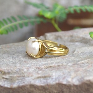 May include: A gold ring featuring a large white pearl set in the centre. The ring has a simple design with a band that is slightly wider at the front.