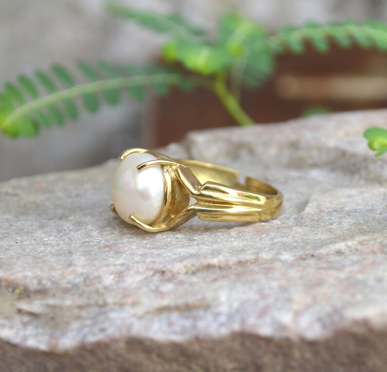May include: A gold ring featuring a large white pearl set in the centre. The ring has a simple design with a band that is slightly wider at the front.
