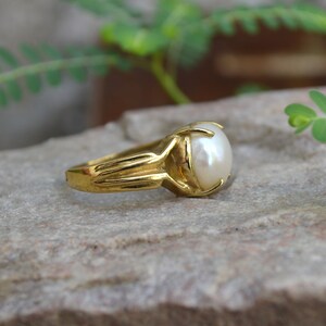 May include: A gold ring with a large, white pearl set in a prong setting. The ring is on a grey, textured surface.