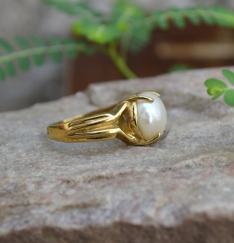 May include: A gold ring with a large, white pearl set in a prong setting. The ring is on a grey, textured surface.