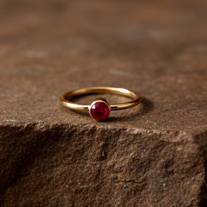 May include: A gold ring featuring a round, deep red gemstone. The ring has a slender, polished band, and the gemstone is set in a simple bezel. The ring is presented on a textured, brown surface.