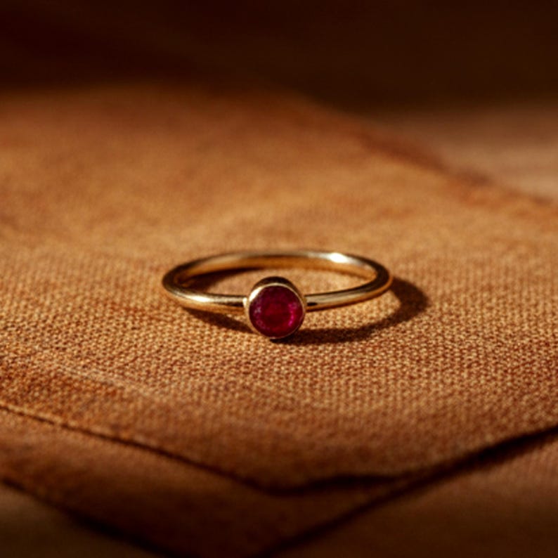 May include: A gold ring featuring a small, circular, deep red gemstone. The ring rests on a textured, brown fabric, contrasting the shiny metal with the matte background. The ring's band is slender and delicate.