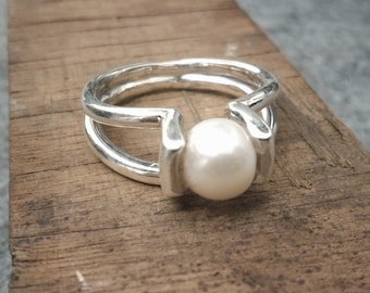 May include: A silver ring featuring a large, round, white pearl. The ring has a distinctive design with a double band, cradling the pearl in the centre. The ring is shown on a wooden surface.