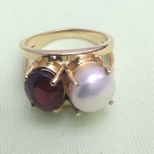 Può includere: Un anello d'oro con una pietra rossa e una perla bianca. L'anello è incastonato su una fascia semplice.