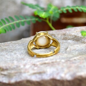 May include: A gold ring with a white oval-shaped stone set in the centre. The ring is adjustable and has a simple design.