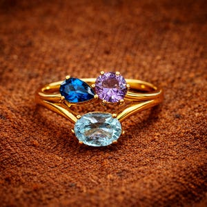 May include: Three gold rings with gemstone settings. The top ring features a blue pear-shaped stone and a purple round stone. The bottom ring has a light blue oval stone. The rings are set against a brown textured background.