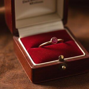 May include: A gold ring with a round, red gemstone sits in an open wooden jewellery box with a red velvet interior. The ring is the focal point, and the box is brown with a brass clasp. The ring's design is highlighted by the box's presentation.