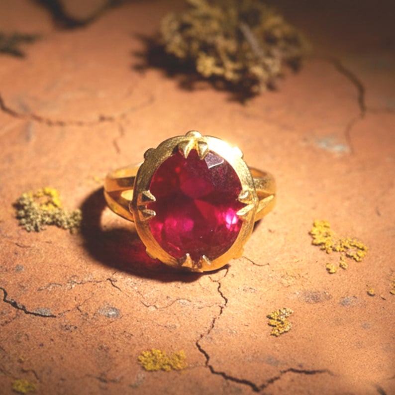 May include: A gold ring with a large, oval-cut, ruby-red gemstone. The ring's setting has a decorative, scalloped design. The ring is set against a textured, brown surface with small patches of yellow and green.