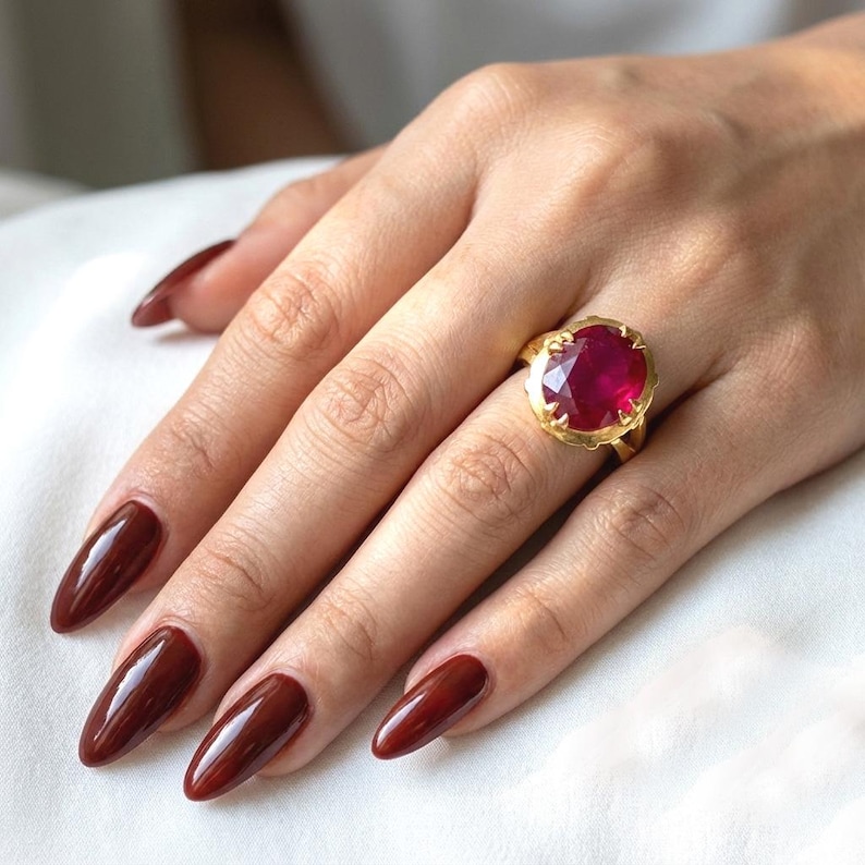 May include: A gold ring featuring a large, faceted, oval-cut ruby gemstone. The ring has a detailed, ornate setting. The hand has long, polished, burgundy-colored nails. The background is a soft, white fabric.