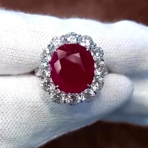 May include: A large, oval-shaped ruby ring set in a halo of sparkling diamonds. The ruby is a deep red color and is surrounded by a ring of smaller diamonds.