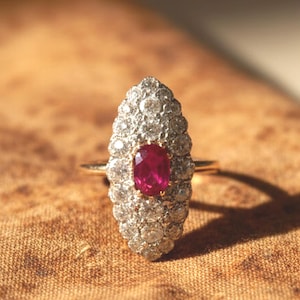 Peut inclure: Une bague de style vintage avec un rubis ovale central, entouré d'un groupe de petits diamants étincelants. La bague a un anneau en or et est posée sur un fond brun texturé. Le rubis est d'un rouge profond.