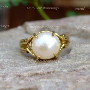 May include: A gold ring featuring a large, white pearl set in the centre. The ring has a delicate, intricate design.