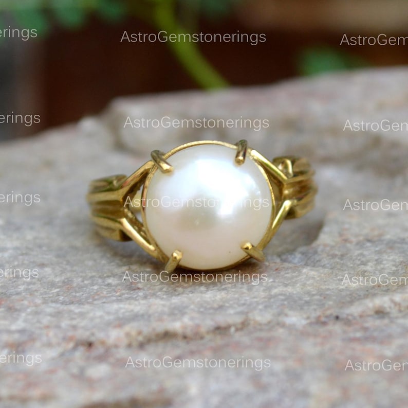 May include: A gold ring featuring a large, white pearl set in the centre. The ring has a delicate, intricate design.