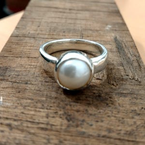 Puede incluir: Un anillo de plata con una perla blanca y redonda grande en el centro. El anillo tiene una banda gruesa y pulida, y la perla es el punto focal, sobre una superficie de madera.