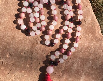 May include: A mala or prayer bead necklace with a red tassel. The beads are made of white, pink, and dark red stones.