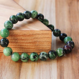 May include: A beaded bracelet featuring round, polished beads in shades of green and black. The bracelet is strung together, creating a flexible band. The beads have a marbled appearance, with variations in color and texture.