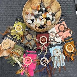 May include: A collection of ten small, colorful pouches with zipper closures. Each pouch features a small dreamcatcher with feathers. The pouches are arranged in a circle on a black surface. A wooden bowl filled with colorful stones is in the background.