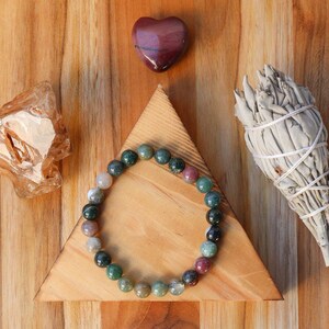 May include: A green, brown, and pink gemstone bracelet on a wooden triangle with a heart-shaped stone, a clear crystal, and a bundle of white sage.