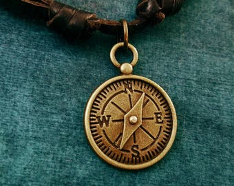 May include: A brown leather cord necklace with a brass compass pendant. The compass features a pointed arrow and the letters N, S, E, and W.