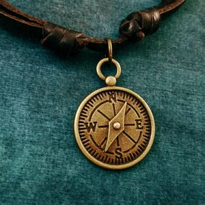 May include: A brown leather cord necklace with a brass compass pendant. The compass features a pointed arrow and the letters N, S, E, and W.