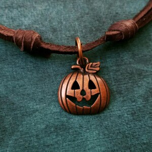 May include: A brown leather cord necklace with a copper-colored pumpkin charm. The pumpkin has a carved jack-o'-lantern face.
