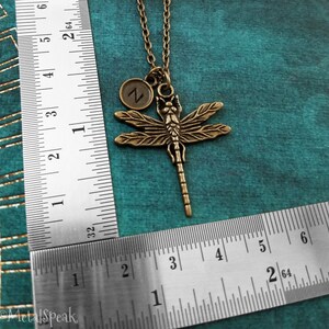 May include: A bronze dragonfly pendant necklace with a small initial charm. The charm is a circle with the letter "Z" in the center. The necklace is on a chain and is measured against a ruler.