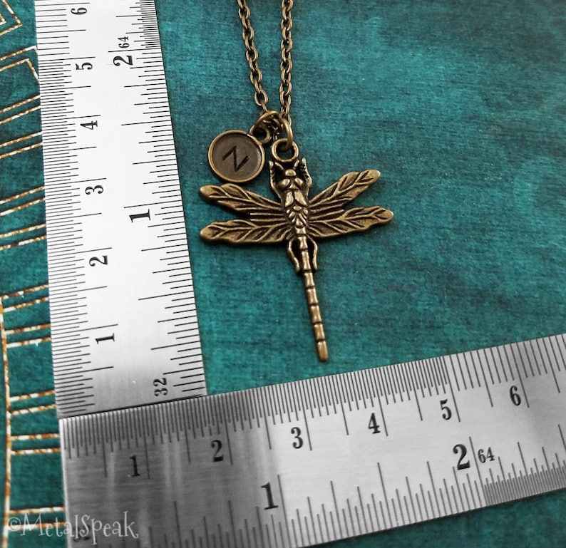 May include: A bronze dragonfly pendant necklace with a small initial charm. The charm is a circle with the letter "Z" in the center. The necklace is on a chain and is measured against a ruler.