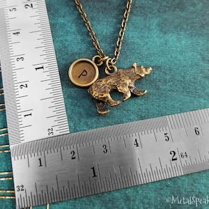 May include: A gold-toned necklace with a bear charm and a round charm with the letter "P" engraved on it. The necklace is measured against a ruler.