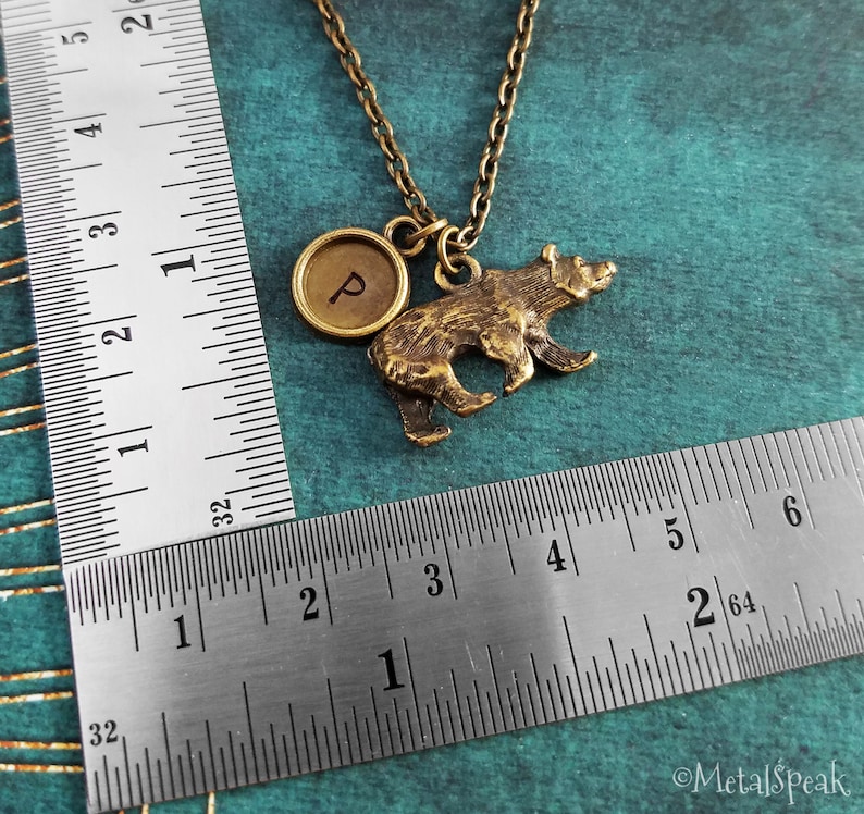 May include: A gold-toned necklace with a bear charm and a round charm with the letter "P" engraved on it. The necklace is measured against a ruler.