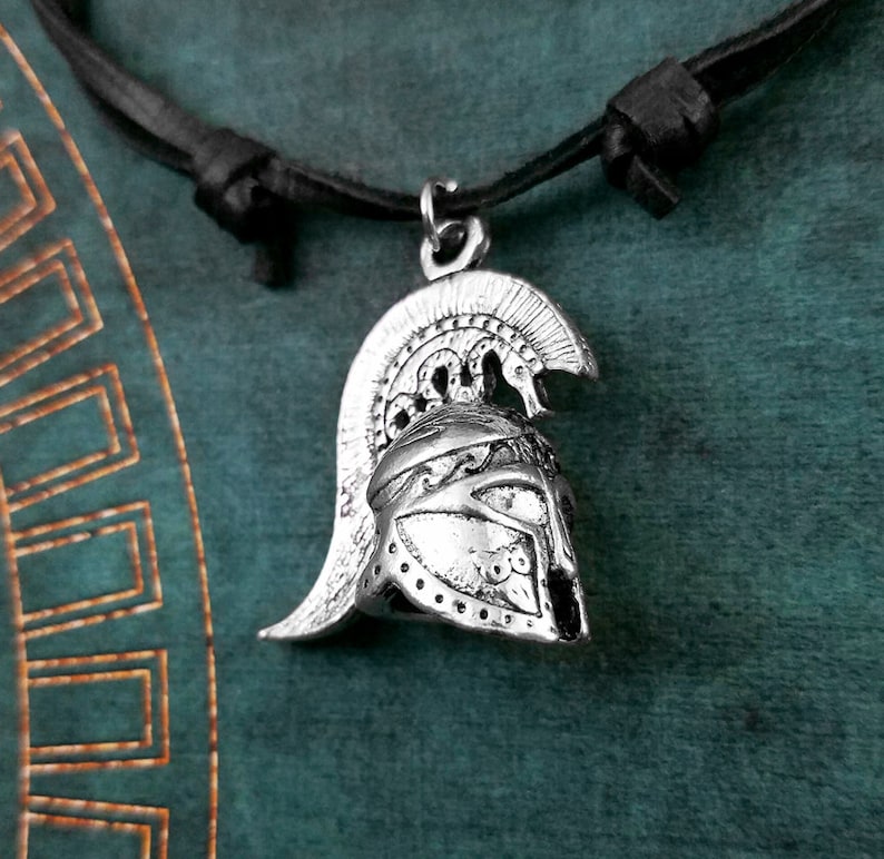 May include: A silver-toned pendant necklace featuring a detailed depiction of a Greek warrior helmet. The necklace is on a black leather cord.