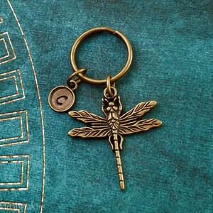 May include: A bronze dragonfly keychain with a letter 'C' charm. The keychain is on a teal background.