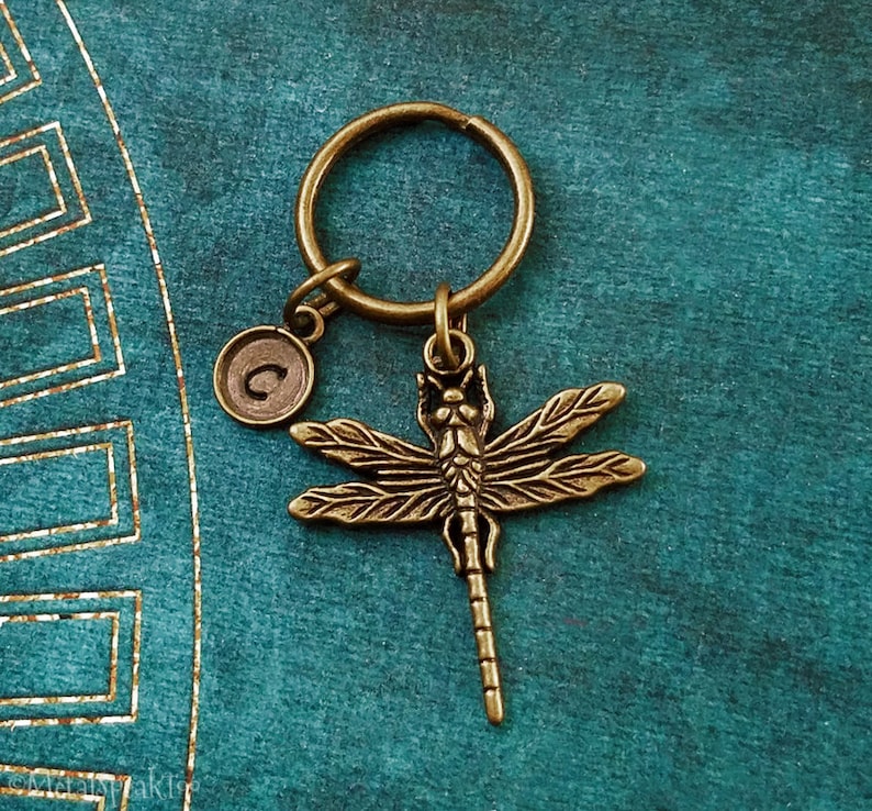 May include: A bronze dragonfly keychain with a letter 'C' charm. The keychain is on a teal background.
