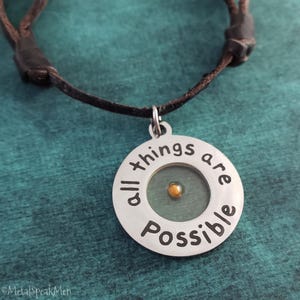 May include: A silver pendant necklace with the words "all things are possible" engraved around the edge. The pendant features a small, gold-coloured seed in the centre, set against a green background. The necklace has a brown leather cord.