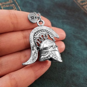 May include: Silver necklace with a detailed pendant of a Greek warrior helmet. The pendant has a textured surface and a raised design. The necklace also features a small silver charm with the letter "R" on it.