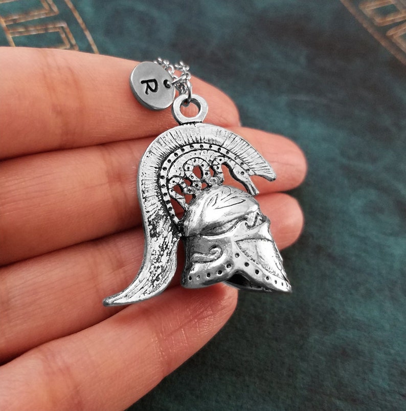 May include: Silver necklace with a detailed pendant of a Greek warrior helmet. The pendant has a textured surface and a raised design. The necklace also features a small silver charm with the letter "R" on it.