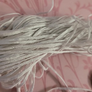 Puede incluir: Un haz de cordón blanco, probablemente de algodón, se muestra sobre un fondo floral rosa. El cordón está bien enrollado y parece nuevo. Una moneda se utiliza como referencia de tamaño.