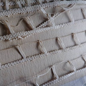 May include: Close-up of a textile with rows of white, looped trim and vertical, looped accents. The background is a light tan color, with visible stitching. The trim has a wavy, decorative edge, creating a textured surface.