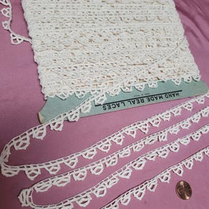 May include: A roll of cream-coloured lace with a decorative edge, and some loose strands. The lace is displayed on a pink surface, with a penny for scale. The packaging reads "HAND MADE REAL LACES".