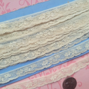 May include: Several rolls of delicate, cream-colored lace trim are displayed on a blue surface. The lace features a scalloped edge and intricate floral patterns. A smaller, pinkish-beige lace trim is also visible, along with a dark brown coin.