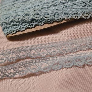 Puede incluir: Un rollo de cinta de encaje azul claro con un delicado diseño floral. Una longitud de la cinta está desenrollada, junto a una moneda para comparar el tamaño. El encaje está sobre un fondo de tela rosa.