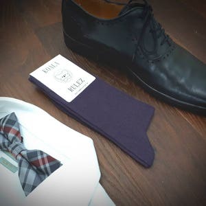 May include: A pair of dark purple socks with a white tag that says "Koala Rulez". The socks are lying on a wooden surface next to a black leather shoe and a white shirt with a plaid bow tie.