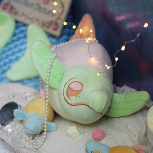 May include: A plush green and white fish toy with a pearl necklace. The fish has a closed eye and a pink and orange nose. The fish is lying on a white surface with other plush toys.