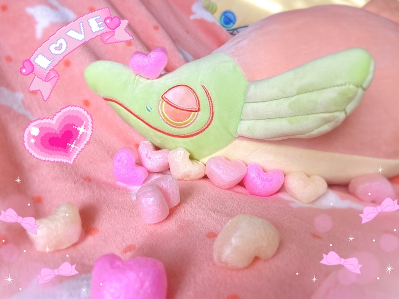 May include: A plush green and yellow dragon with wings lying on a pink blanket with the word "LOVE" in pink letters on a banner above it.  There are many pink heart-shaped objects scattered around the dragon.