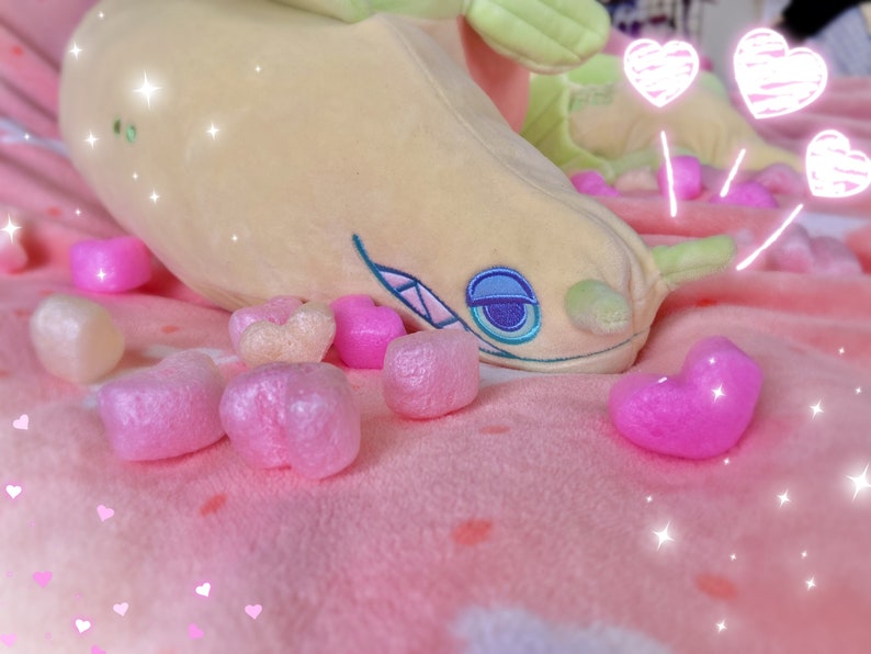 A soft pastel green moray eel plush with blue eyes and a toothy smile. It's laying on a pink blanket, surrounded by hearts and sparkles.