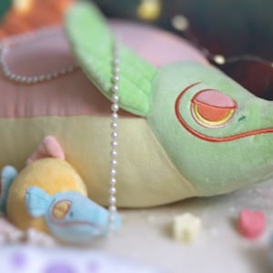 May include: A green and pink plush toy shaped like a fish with a pearl necklace. The fish is lying on a white surface with a smaller yellow and blue plush fish toy beside it.