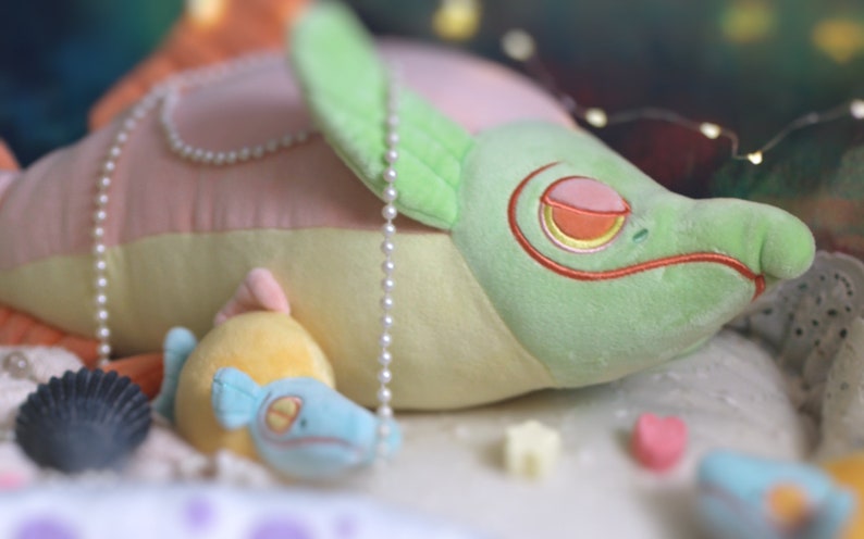 May include: A green and pink plush toy shaped like a fish with a pearl necklace. The fish is lying on a white surface with a smaller yellow and blue plush fish toy beside it.