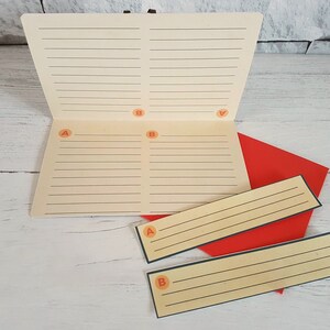May include: A white notebook with lined pages divided into four sections labeled A and B. The notebook is open and lying on a white wooden surface. There are two smaller lined cards with A and B labels, also on the surface.